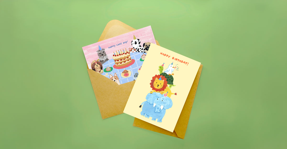 Two illustrated birthday cards with brown envelopes on a solid green background. One card shows a pink background with animal characters around a birthday cake, and the other shows a yellow background with a lion and blue elephant under the text 'HAPPY BIRTHDAY!'.